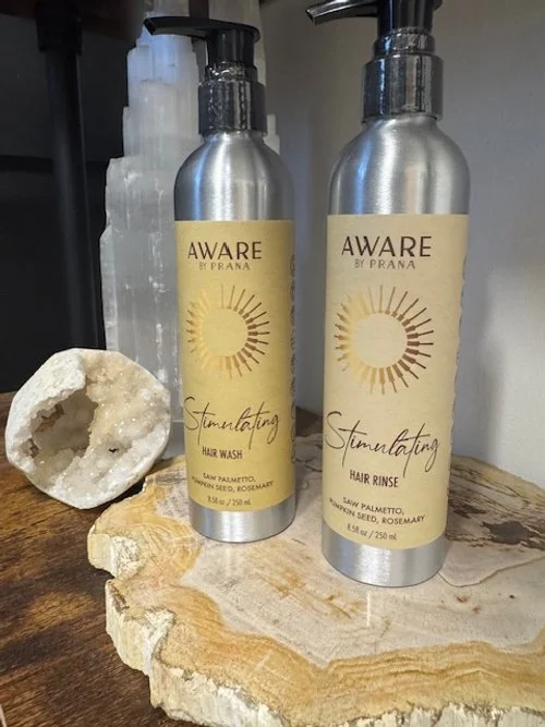 Two bottles of Aware by Prana hair wash and hair rinse on a wooden surface, with a large salt crystal and a decorative mineral or rock in the background.