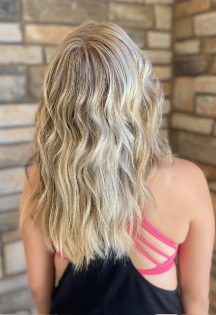 Back view of a woman with long, wavy blonde hair wearing a pink strappy top and a black dress, standing in front of a brick wall.