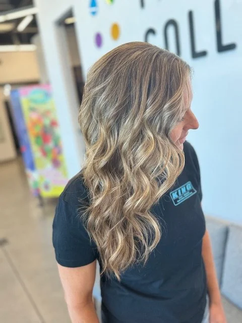 Woman with wavy blonde hair standing indoors at a classroom or office setting.