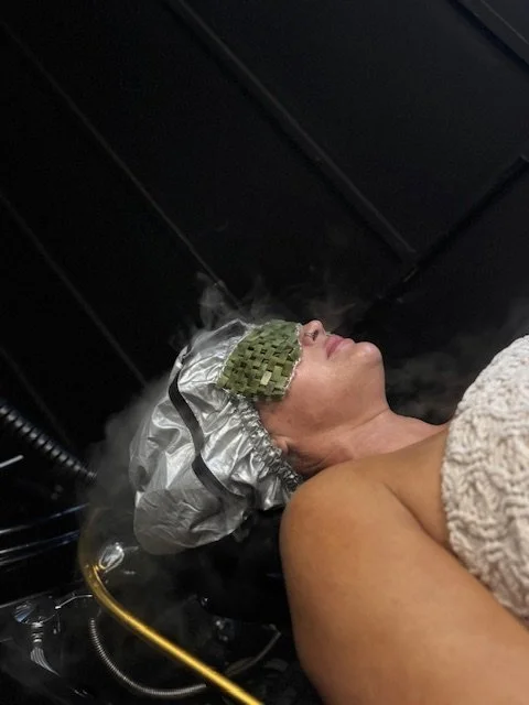 Person lying down with a heat or steam cap on their forehead, connected to a machine, in a dark setting.