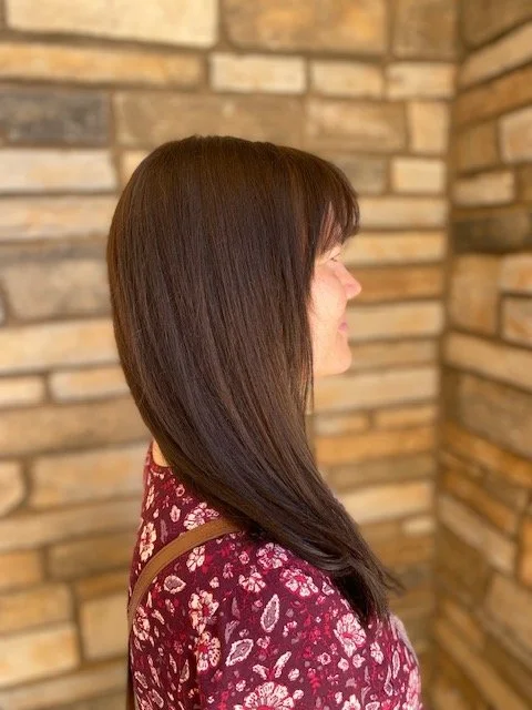 Profile of a woman with long, straight brown hair, wearing a red floral dress, standing against a brick wall.