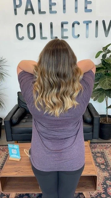 Back of a woman with long, wavy blonde hair standing in front of a wall with the words "Palette Collective", arms raised behind her head. She is wearing a purple shirt and black pants, in a room with a black sofa, a wooden coffee table, and a potted 