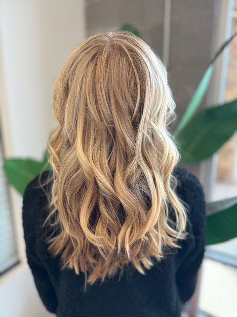 Back view of a woman with long, wavy blonde hair in an indoor setting with plants in the background.