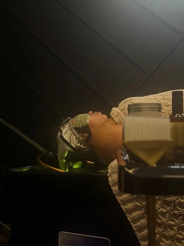 A person lying down wearing a shower cap, possibly preparing for a beauty treatment, with a jar and other items on a table nearby.