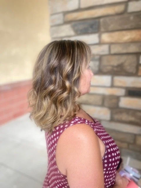 Side view of a woman with shoulder-length wavy blonde hair, wearing a maroon and white polka dot sleeveless top, standing outdoors near a brick wall.