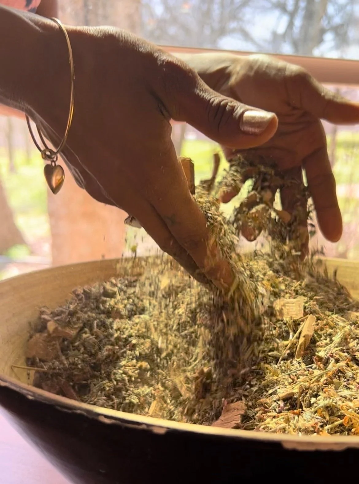 A dark-skinned hand with a gold heart-shaped bracelet stirring a mixture of dried herbs and plant material in a wooden bowl, with a window and trees visible in the background.
