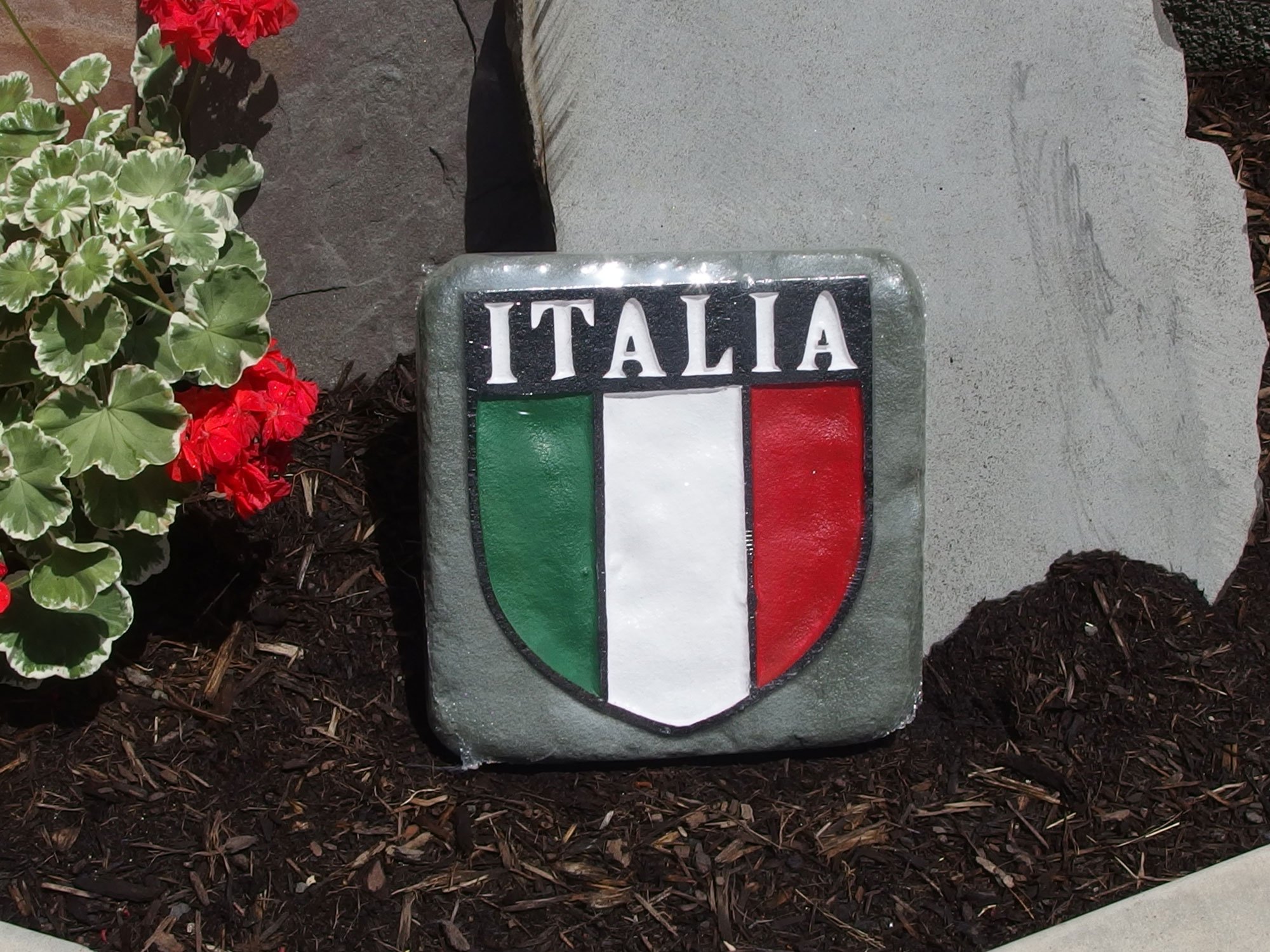 A concrete emblem featuring the Italian flag and the word 'Italia' placed in a flower bed with red and green blooming plants.