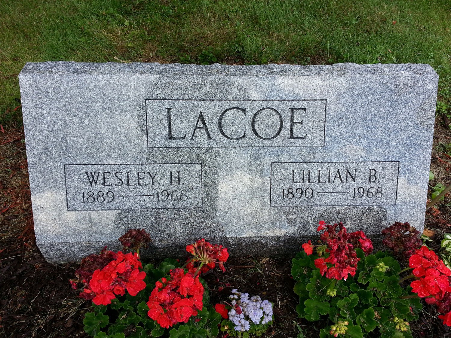 Gray granite headstone with the surname Lacoe engraved at the top, and the names Wesley H. Lacoe (1889-1968) and Lillian B. Lacoe (1890-1968), surrounded by red and white flowers on the ground.