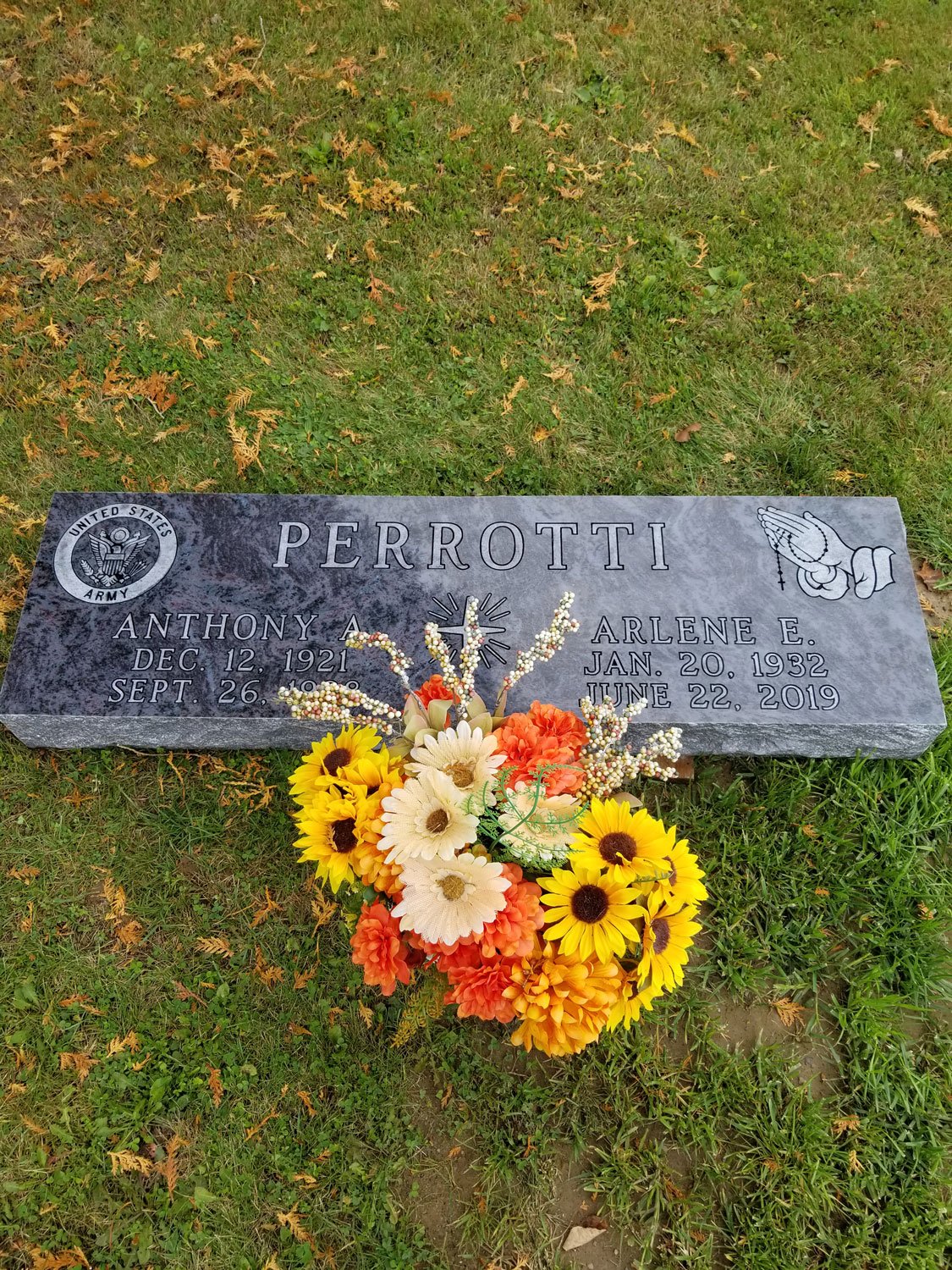 Gravestone with the name Perrotti, surrounded by grass and fallen leaves, decorated with a bouquet of yellow, white, and orange flowers.