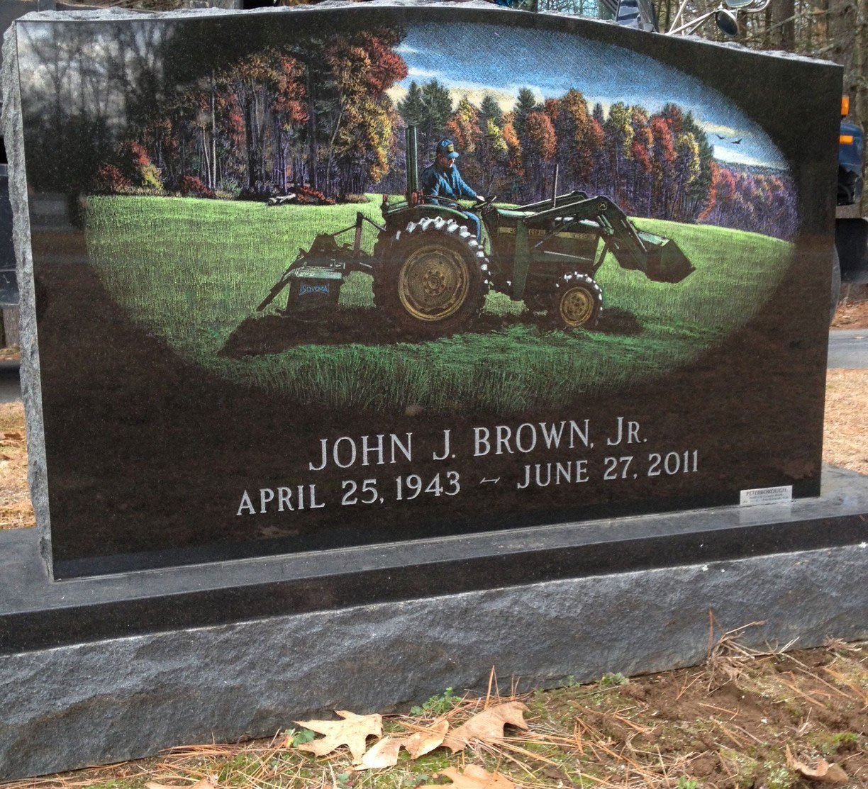 Gravestone with a painted scene of a man driving a tractor on a green field, with trees and a sky in the background, and the inscription "JOHN J. BROWN, JR. APRIL 25, 1943 - JUNE 27, 2011".