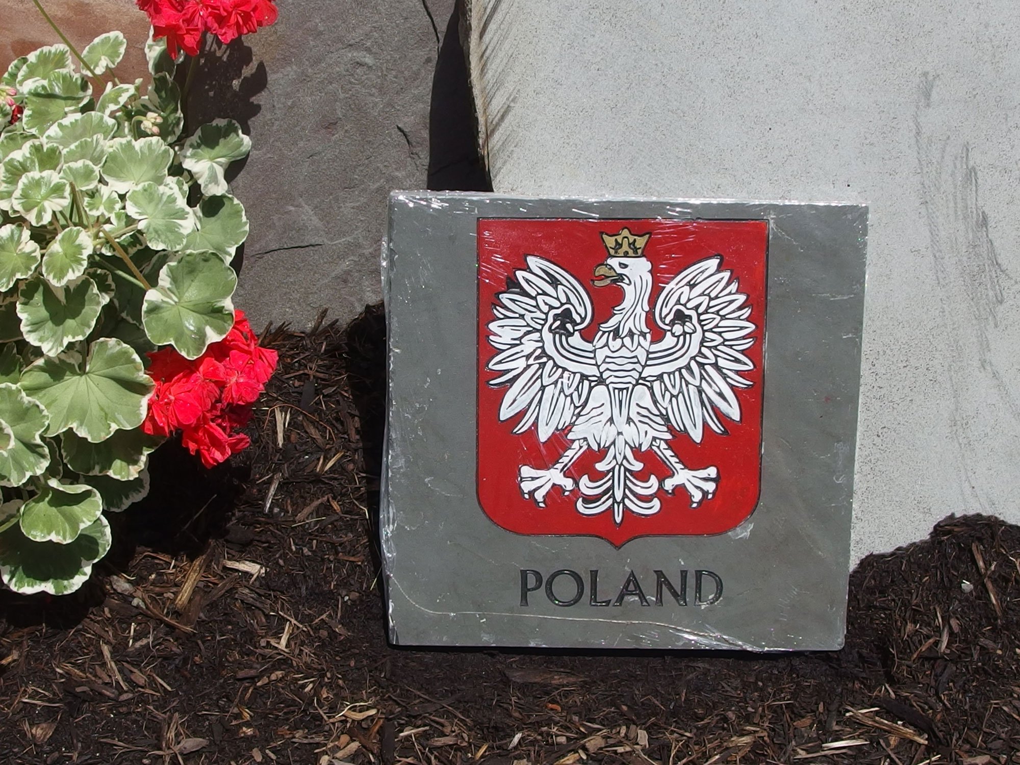 A square plaque with the coat of arms of Poland, featuring a white eagle with a golden crown on a red shield, mounted on a concrete surface, with red and green flowers in the background.