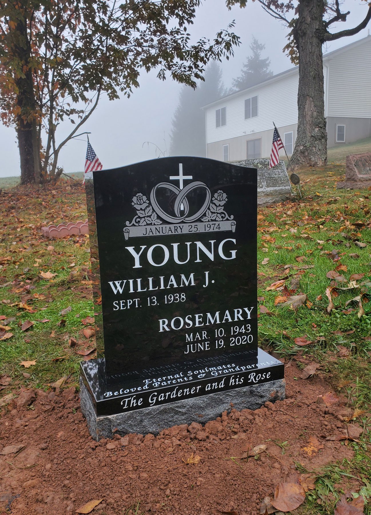 Gravestone for William J. Young and Rosemary with dates, flowers, and American flags on a grassy cemetery hillside with trees and a house in the background