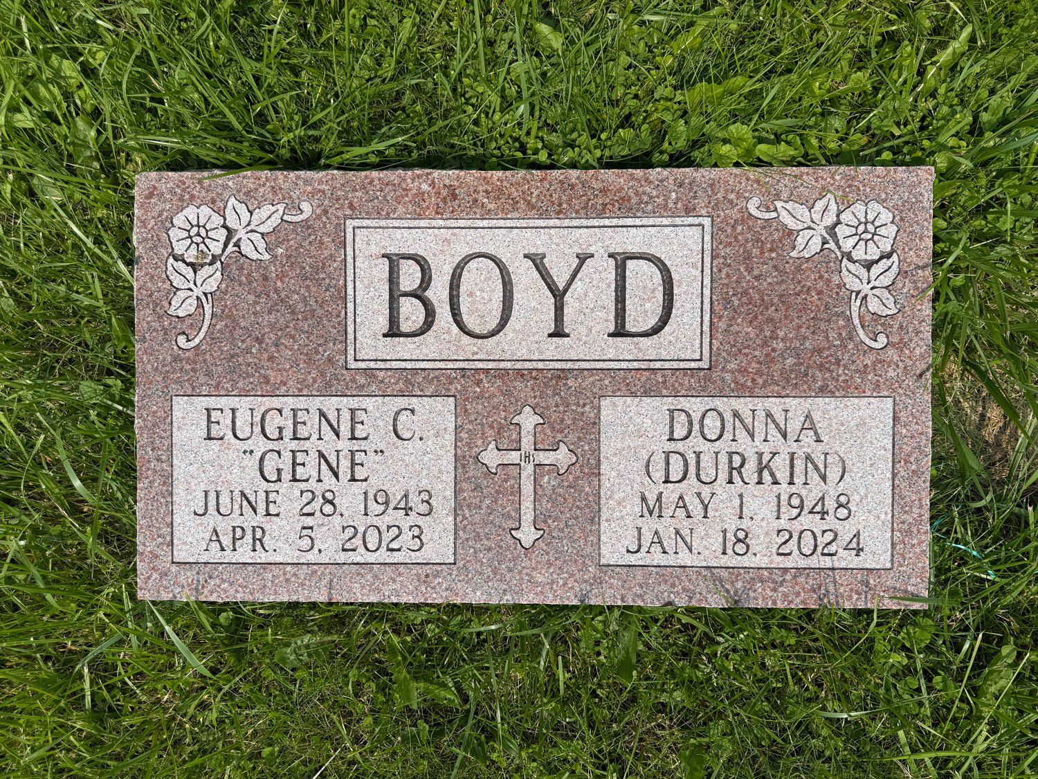 Gravestone for Eugene C. 'Gene' Boyd and Donna (Durkin) Boyd in grass. The gravestone is pinkish granite with floral accents and a cross in the center. Eugene's dates are June 28, 1943 - April 5, 2023, and Donna's are May 1, 1948 - January 18, 2024.