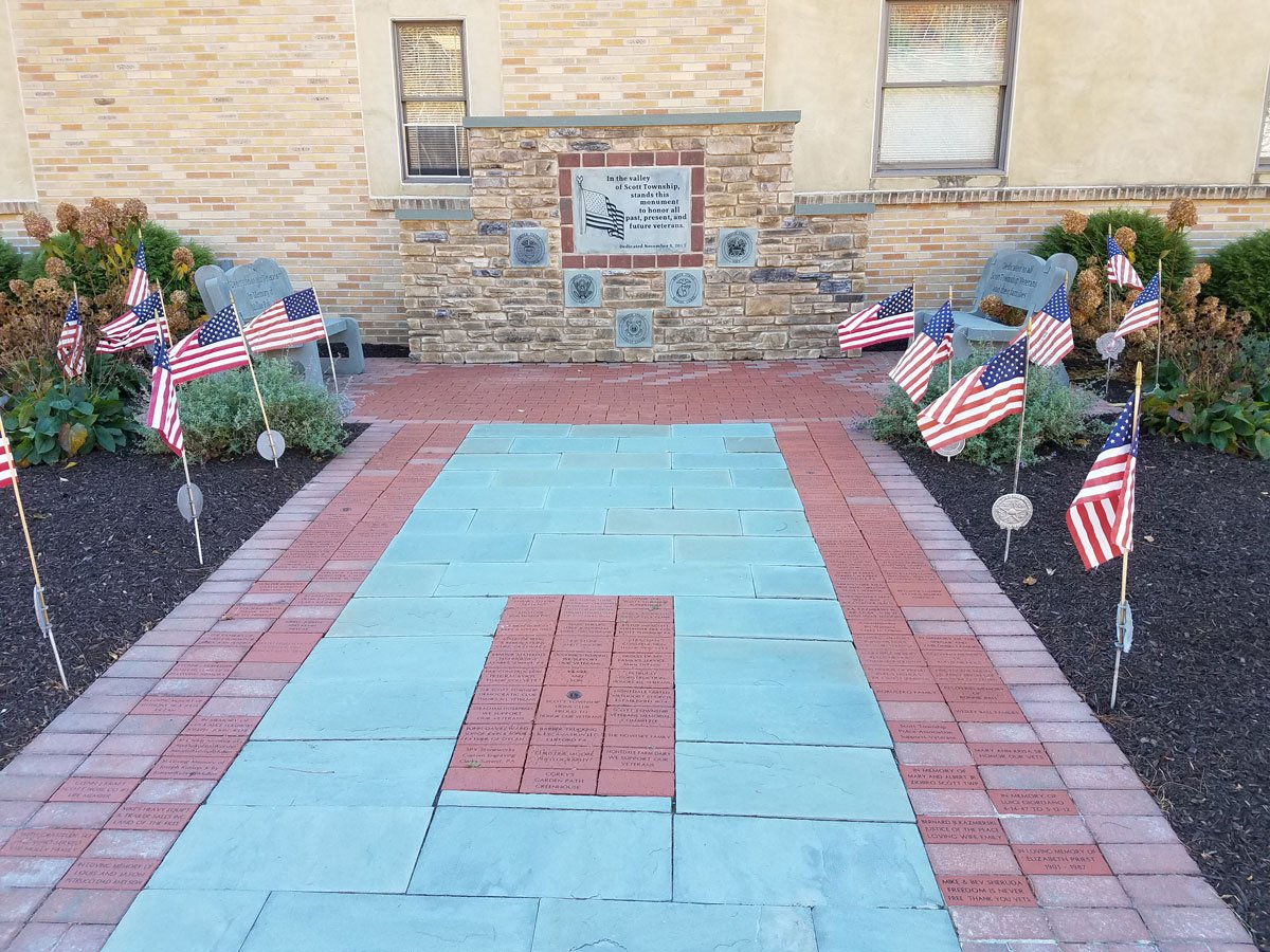 A memorial walkway with American flags on either side, leading to a wall with a plaque honoring veterans, with flowers and shrubs around the area.