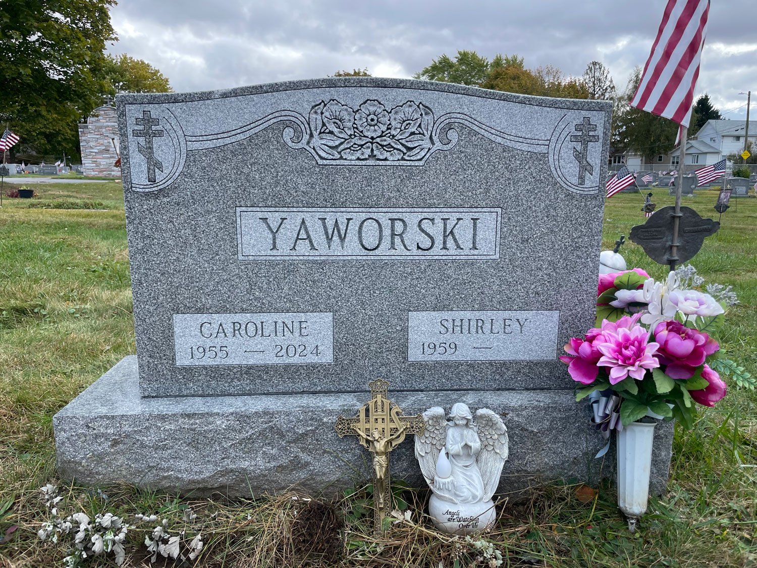 Gravestone with the name Yaworski, marking the resting place of Caroline (1955-2024) and Shirley (1959-). Decorated with American flags, a small angel statue, and religious symbols, in a cemetery with other headstones and trees in the background.