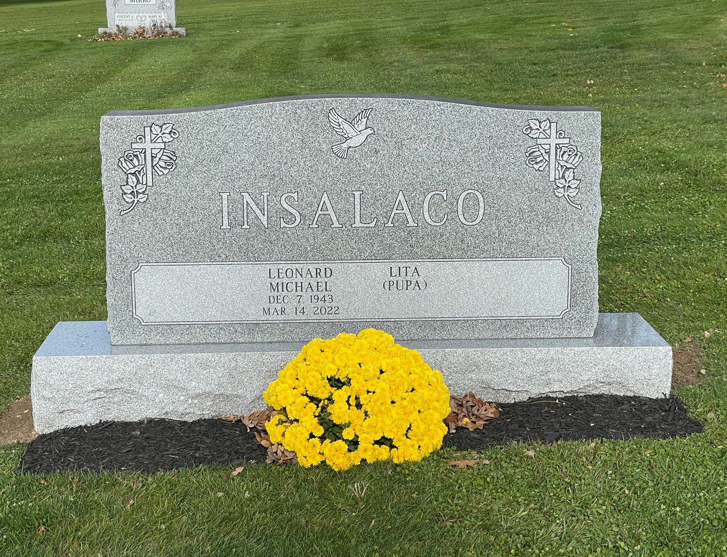 Gray granite gravestone with the name 'Insalaco' engraved, decorated with floral carvings and a dove. The gravestone has the name Leonard Michael Insalaco, born December 7, 1943, died March 14, 2022, and the nickname Lita (Pupa). There is a bunch of