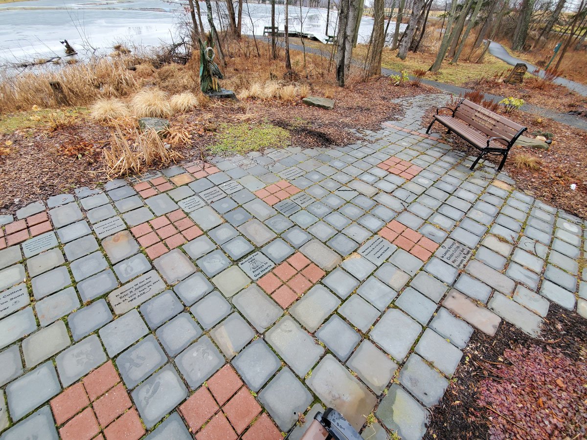 Stone pathway with engraved plaques surrounded by plants, trees, and a lake in a park setting with benches and a walking trail.