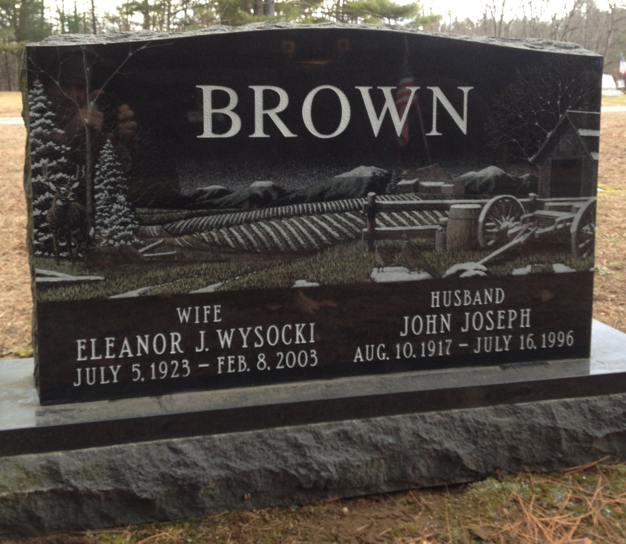 Black headstone engraved with the name 'Brown', depicting a mountain scene, trees, and a wagon in a rural landscape. Names and dates of Eleanor J. Wysocki and John Joseph Brown are inscribed, with details about their birth, death, and relationship.
