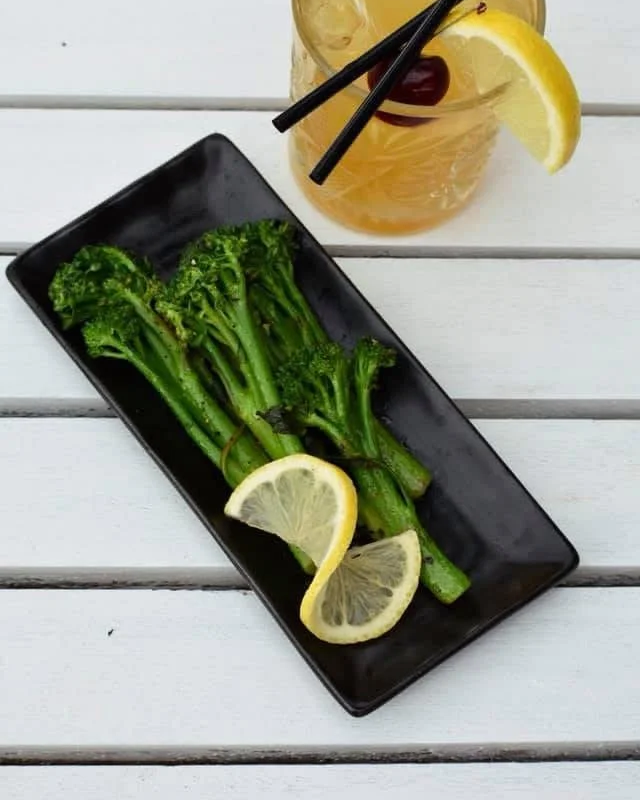 Grilled broccolini with lemon slices on black rectangular plate, side of iced tea with lemon wedge and black straws on white wooden table.