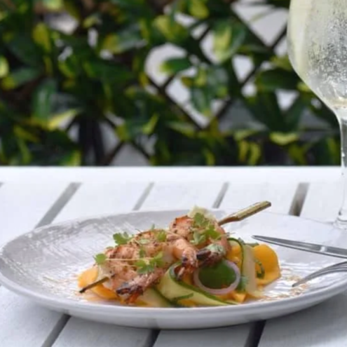 Plate of grilled shrimp with vegetables and herbs, a glass of white wine, and a fork on a white table outside.