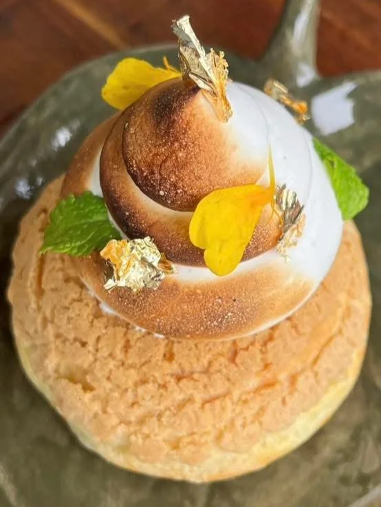 A dessert featuring a round, crumbly cake topped with a glazed chocolate truffle, edible gold leaf, yellow flower petals, a mint leaf, and a glass dome covering the top, served on a plate.