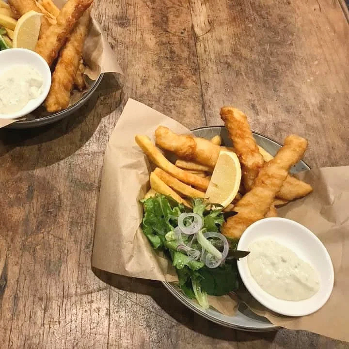Baskets of fried fish fillets with lemon wedges, French fries, a small salad with lettuce and sliced onions, and tartar sauce on a wooden table.