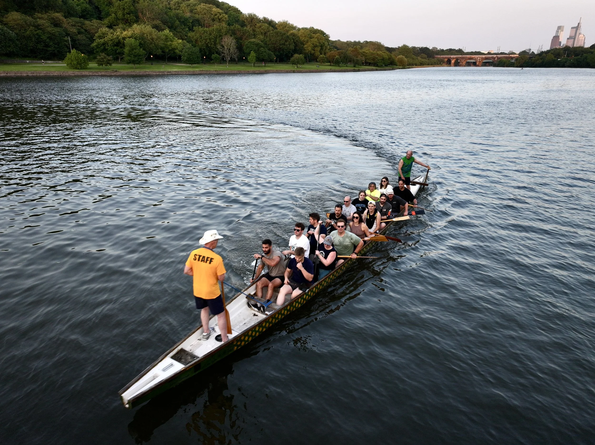 A group of people on a long, narrow boat rowing on a river, guided by a staff member in an orange shirt and white hat, with a green hillside and city buildings in the background.