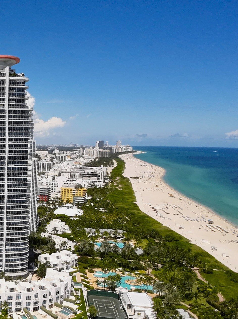 Aerial view of a sunny beach with white sand, turquoise ocean, and high-rise buildings. Green vegetation and swimming pools are visible near the shoreline.