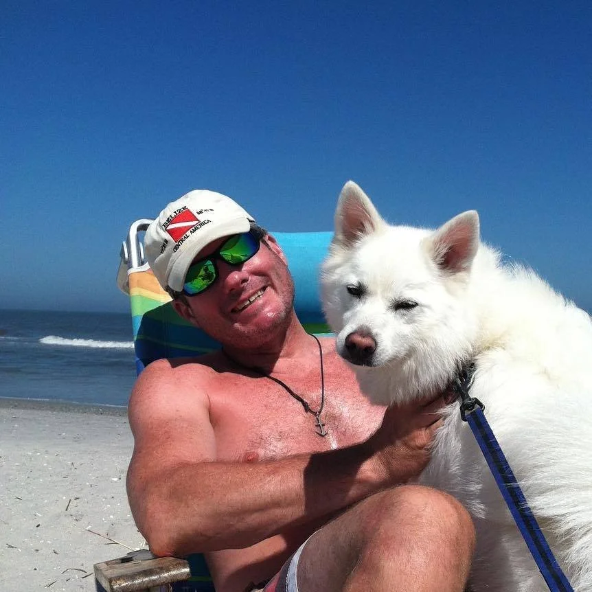 Shirtless man wearing sunglasses and a hat, lying on a beach chair, smiling at the camera, with a white dog on his lap, near the ocean under a clear blue sky.