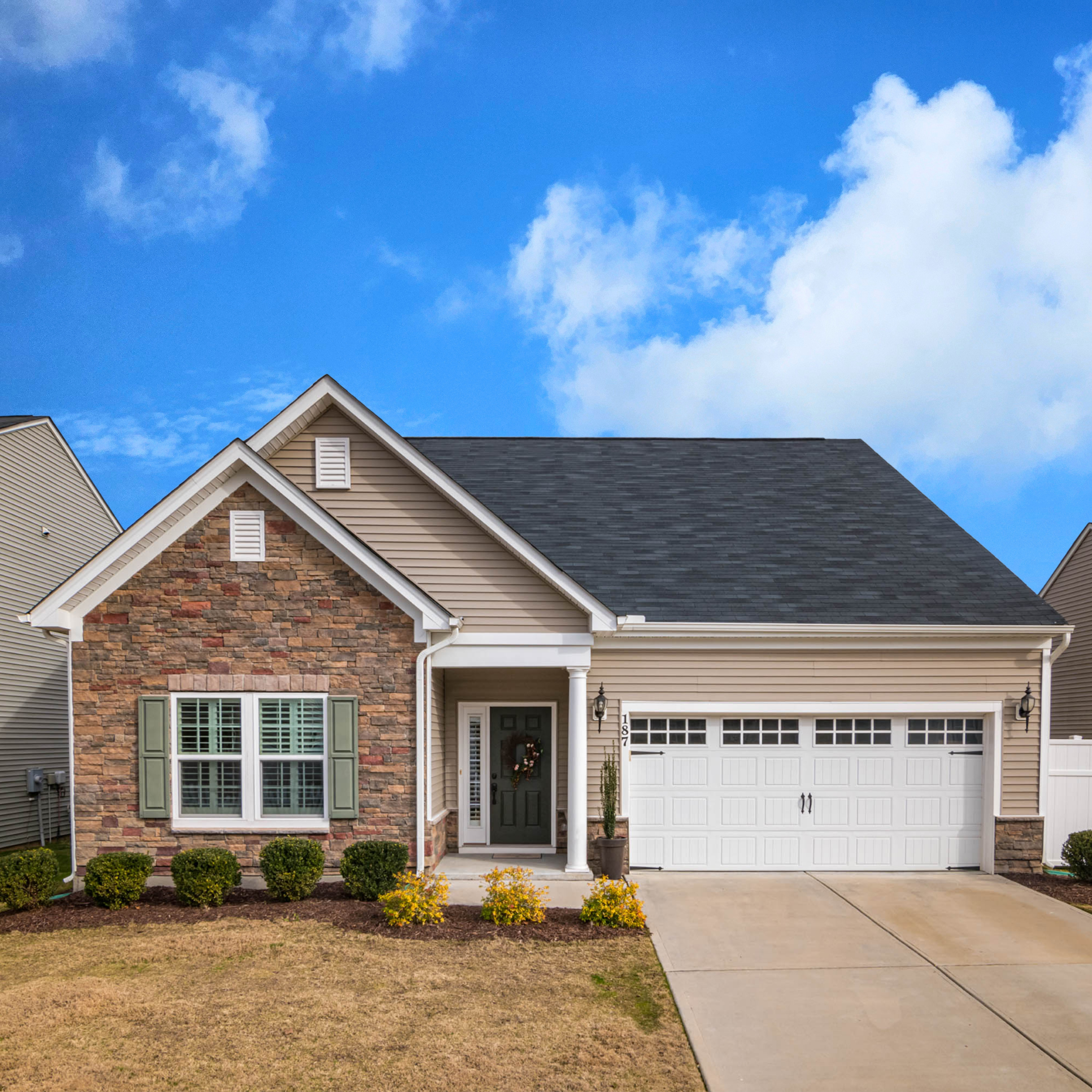 Front exterior of a beige house with a dark gray roof, a black front door with a wreath, a white garage door, and landscaping including small bushes and a pathway.