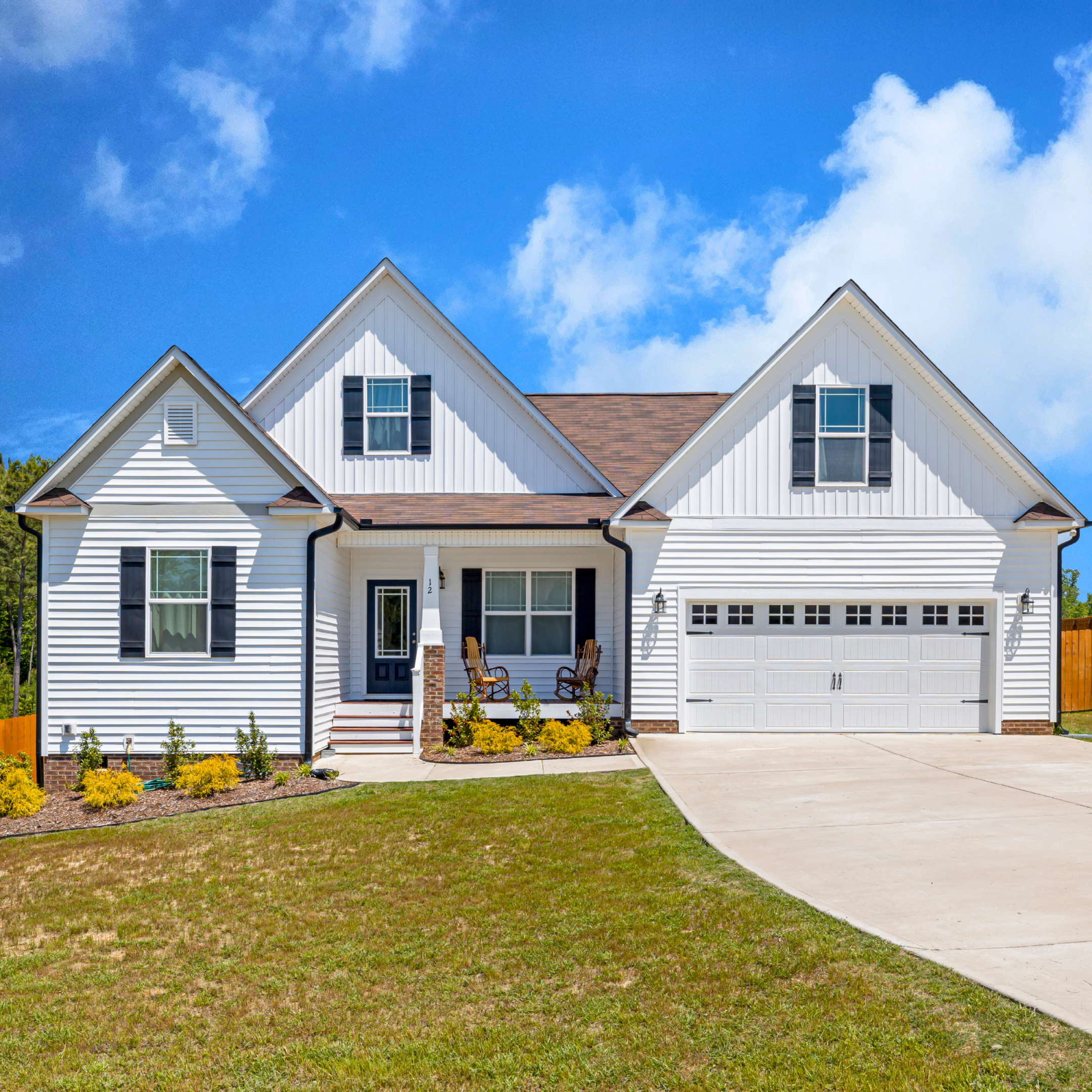 Front view of a white two-story house with black shutters and a brown shingled roof, front yard with grass and plants, concrete sidewalk, and blue sky with clouds.