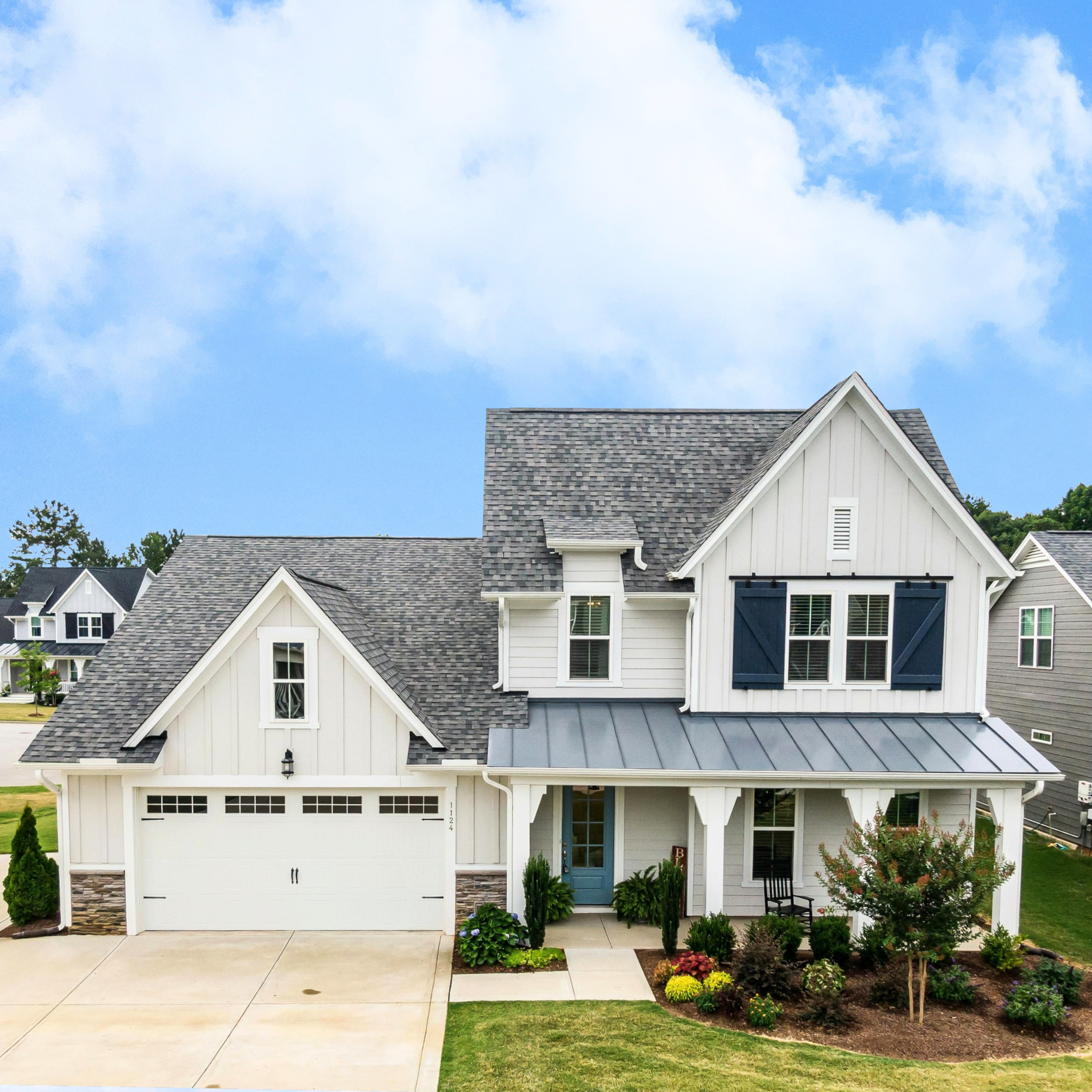 Front view of a modern white house with a gray shingled roof, black shutters, and a blue front door surrounded by landscaped garden and greenery under a blue sky with clouds.