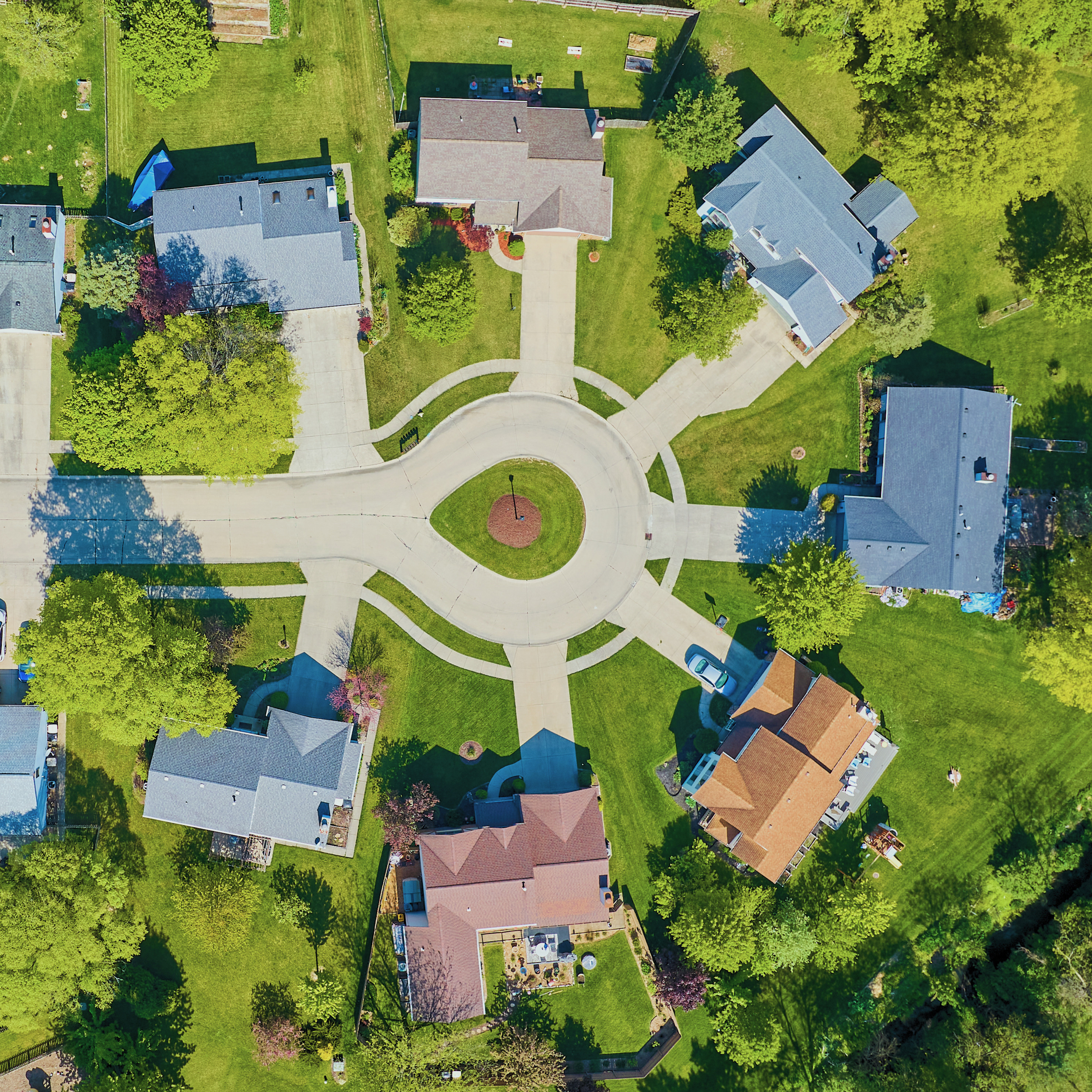 Aerial view of a suburban neighborhood with houses, green lawns, trees, and a circular sidewalk in front of the houses.