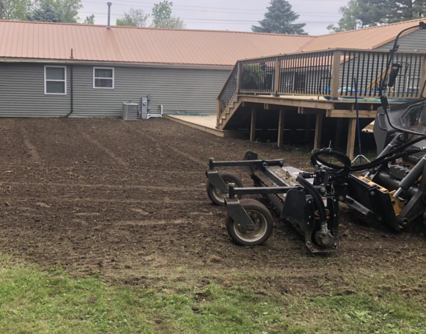 Backyard with a dirt patch, a raised wooden deck with railing, a lawnmower, and a house with gray siding and a red metal roof.