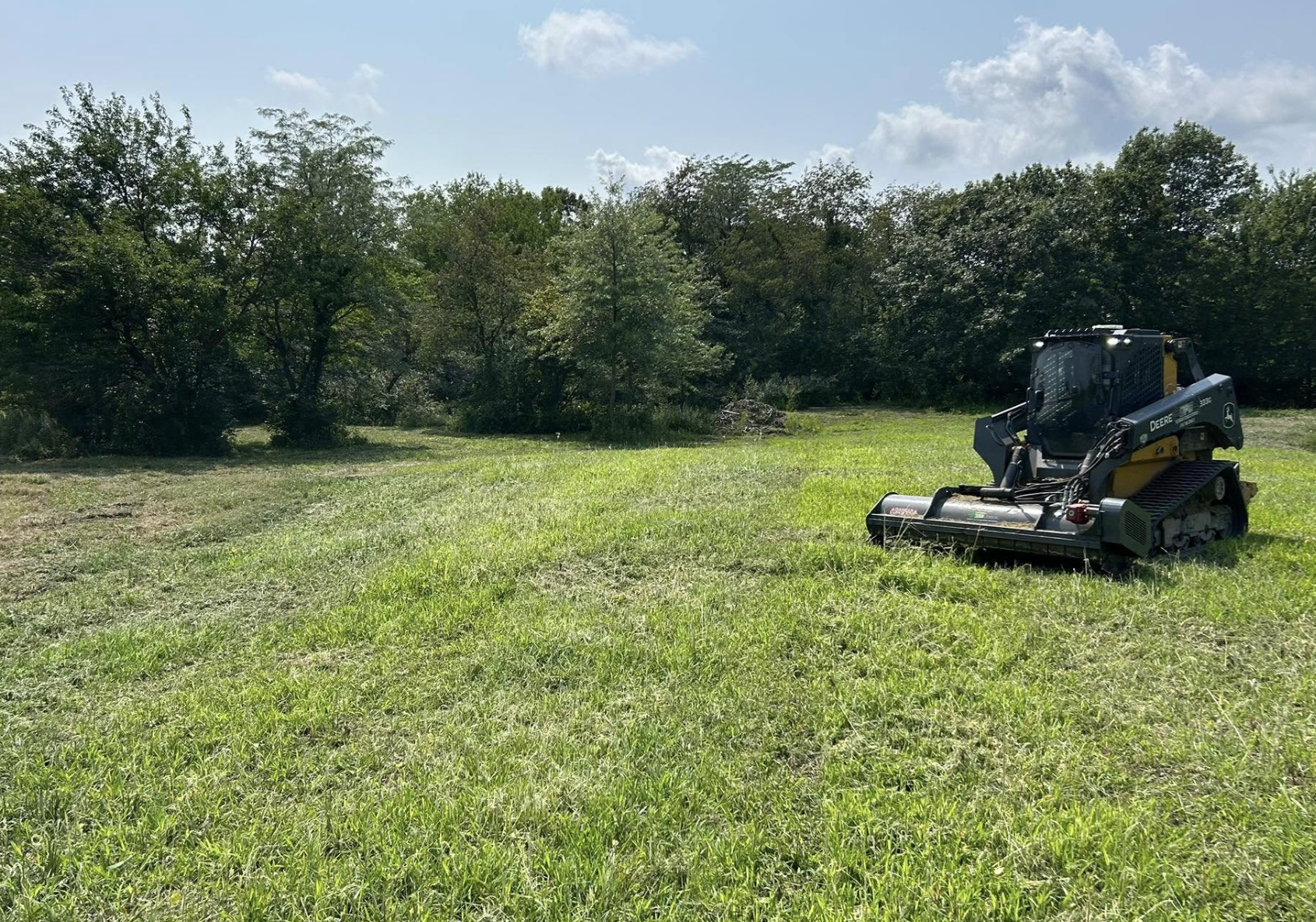 A small tractor on a grassy field with trees in the background and a partly cloudy sky.