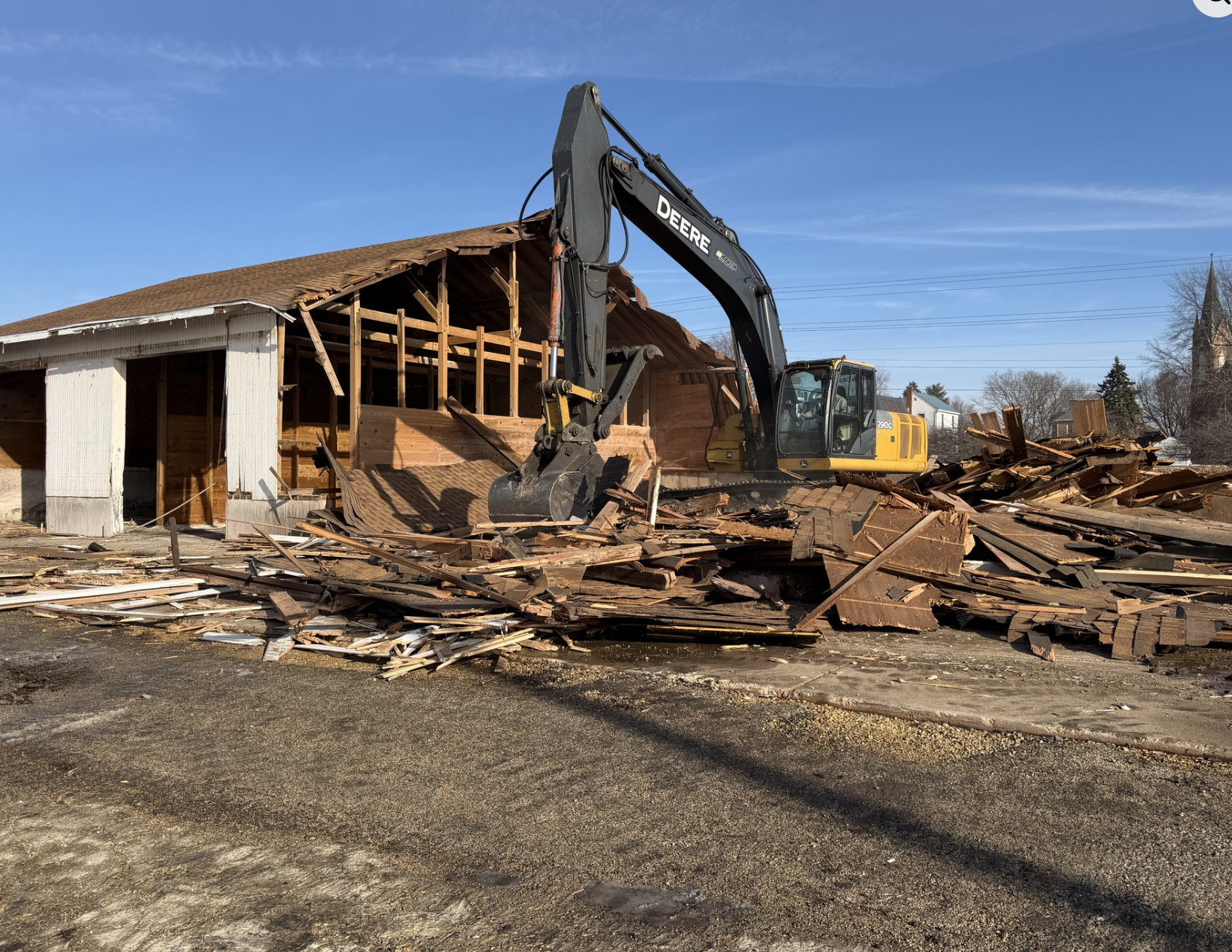 A bulldozer demolishing a wooden house on a clear day with a blue sky. Debris and broken wood scattered on the ground.