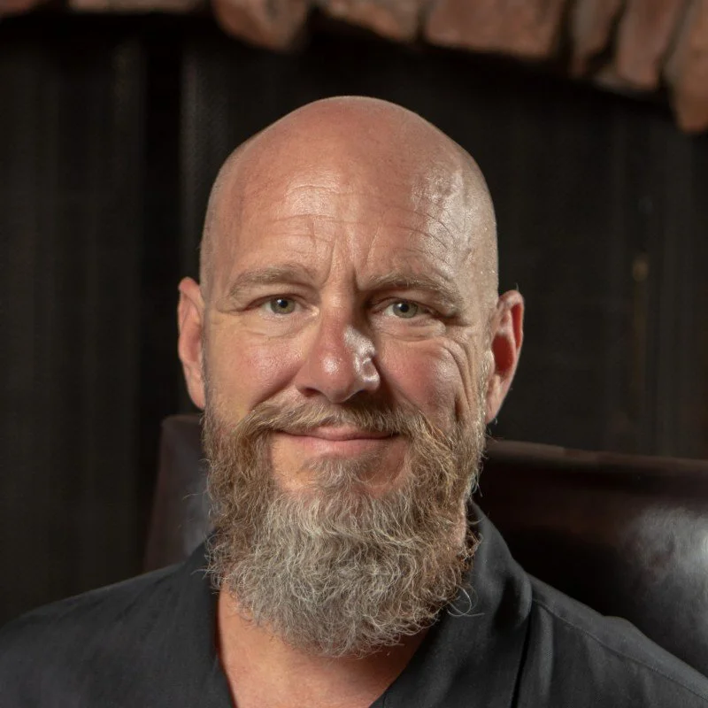 Portrait of a middle-aged man with a bald head and a full gray beard, smiling and wearing a black shirt. The background appears to be a dark interior with a stone or brick wall.