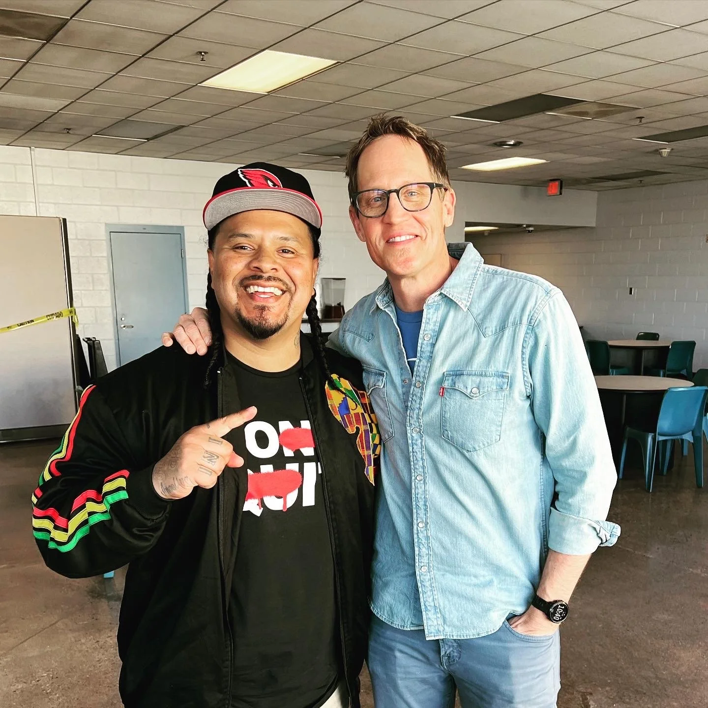 Two men standing together inside a room, smiling at the camera. One man is wearing a black jacket with rainbow stripes on the sleeves, a black t-shirt, and a black cap with a red logo. The other man is wearing glasses, a light blue denim shirt, and light blue pants.