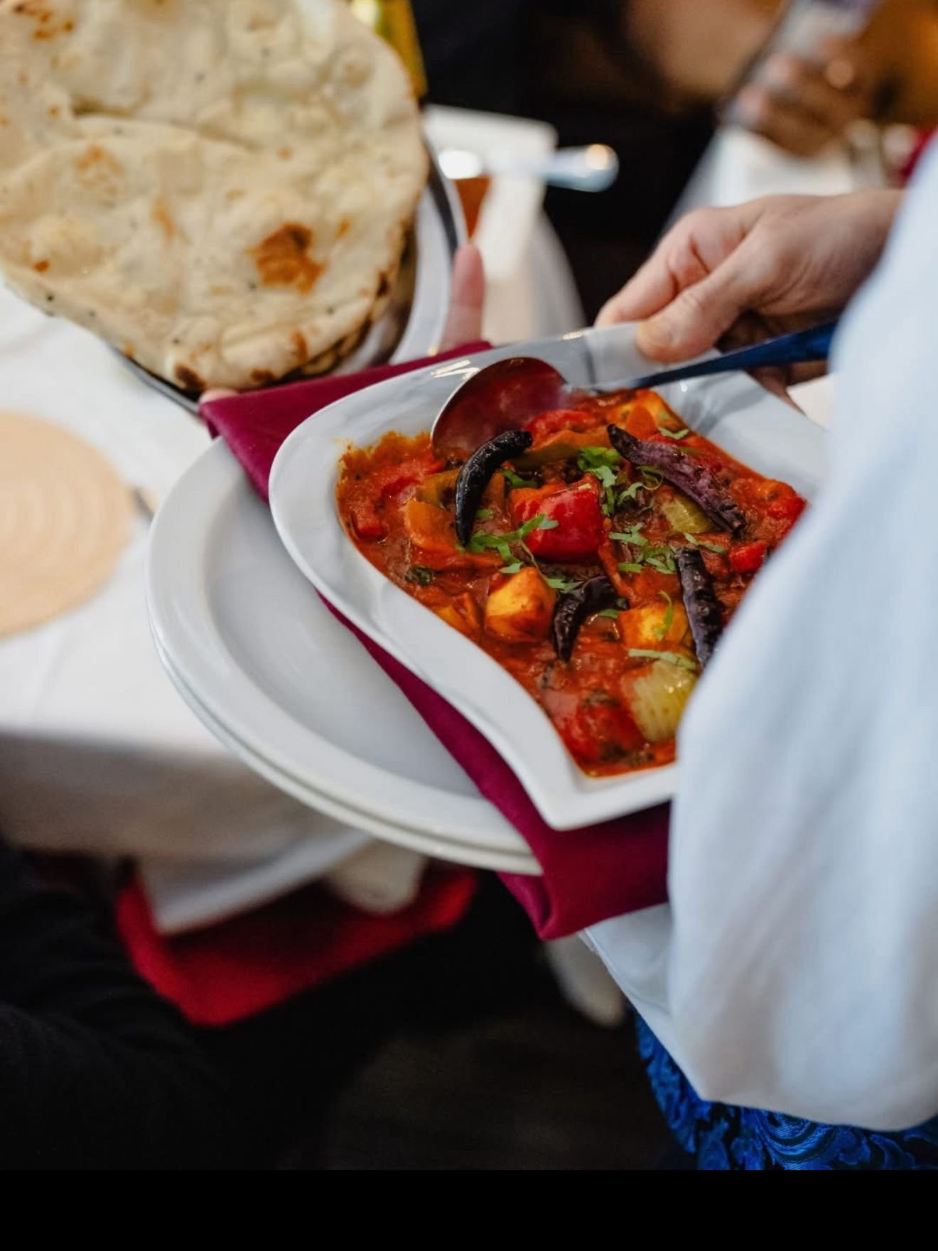 Person holding a plate of vegetable curry with a piece of naan bread in the background.