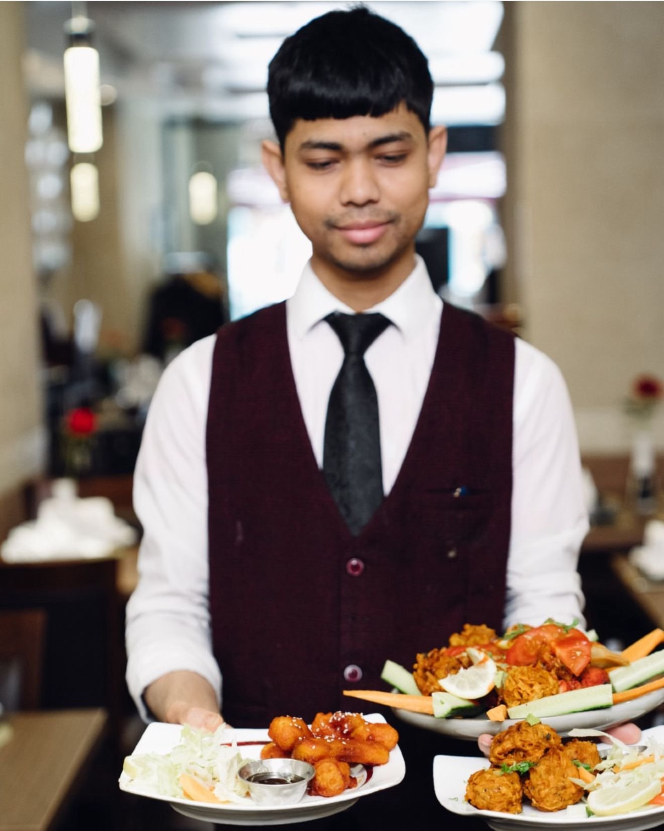 A waiter in a white shirt, black tie, and maroon vest holding two plates of Indian food in a restaurant.