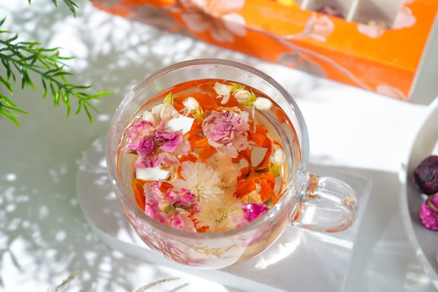 A glass cup filled with water and floating flower petals, including white, pink, and orange blossoms, on a reflective surface with a tissue box in the background.