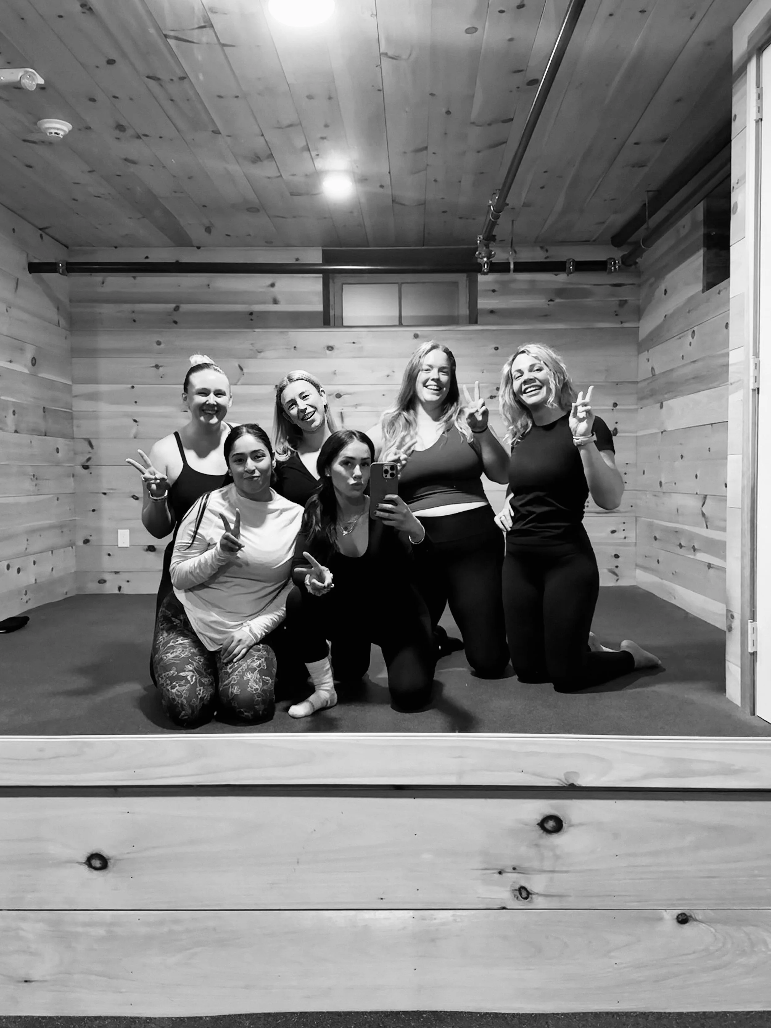 A group of seven women posing for a mirror selfie in a wooden-paneled room, all making peace signs and smiling.
