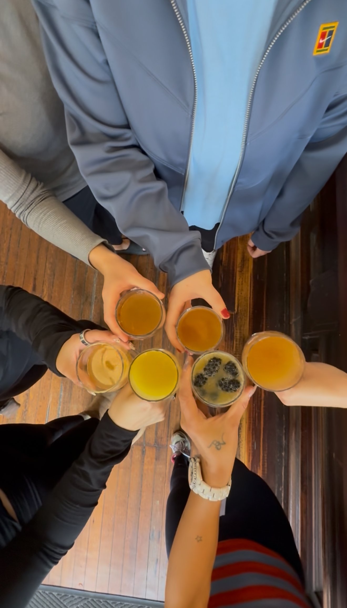 People raising glasses with different drinks in a toast, seen from above, on a wooden table.