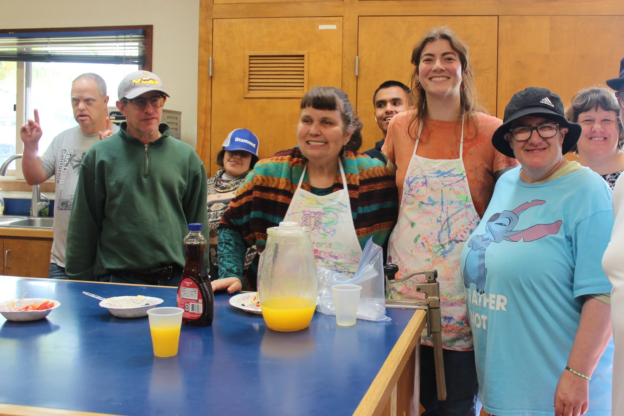 Group of people in a kitchen, smiling and posing for a photo, with breakfast foods and drinks on a counter in front of them.