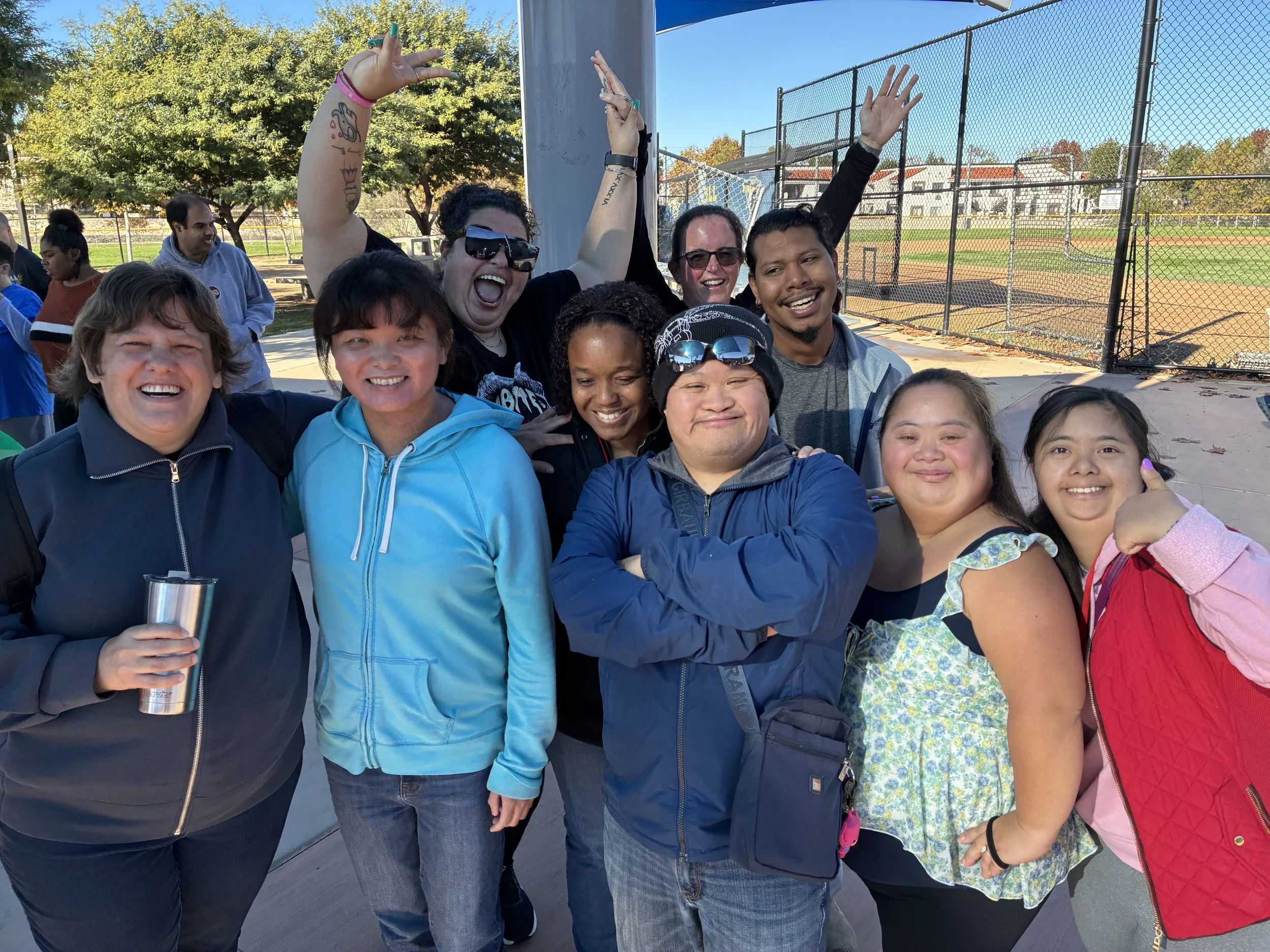 Group of smiling people, including both men and women of various ages and ethnicities, standing outdoors near a tennis court with a blue canopy, trees, and buildings in the background. Some are making energetic hand gestures, and one person is holding a stainless steel tumbler.