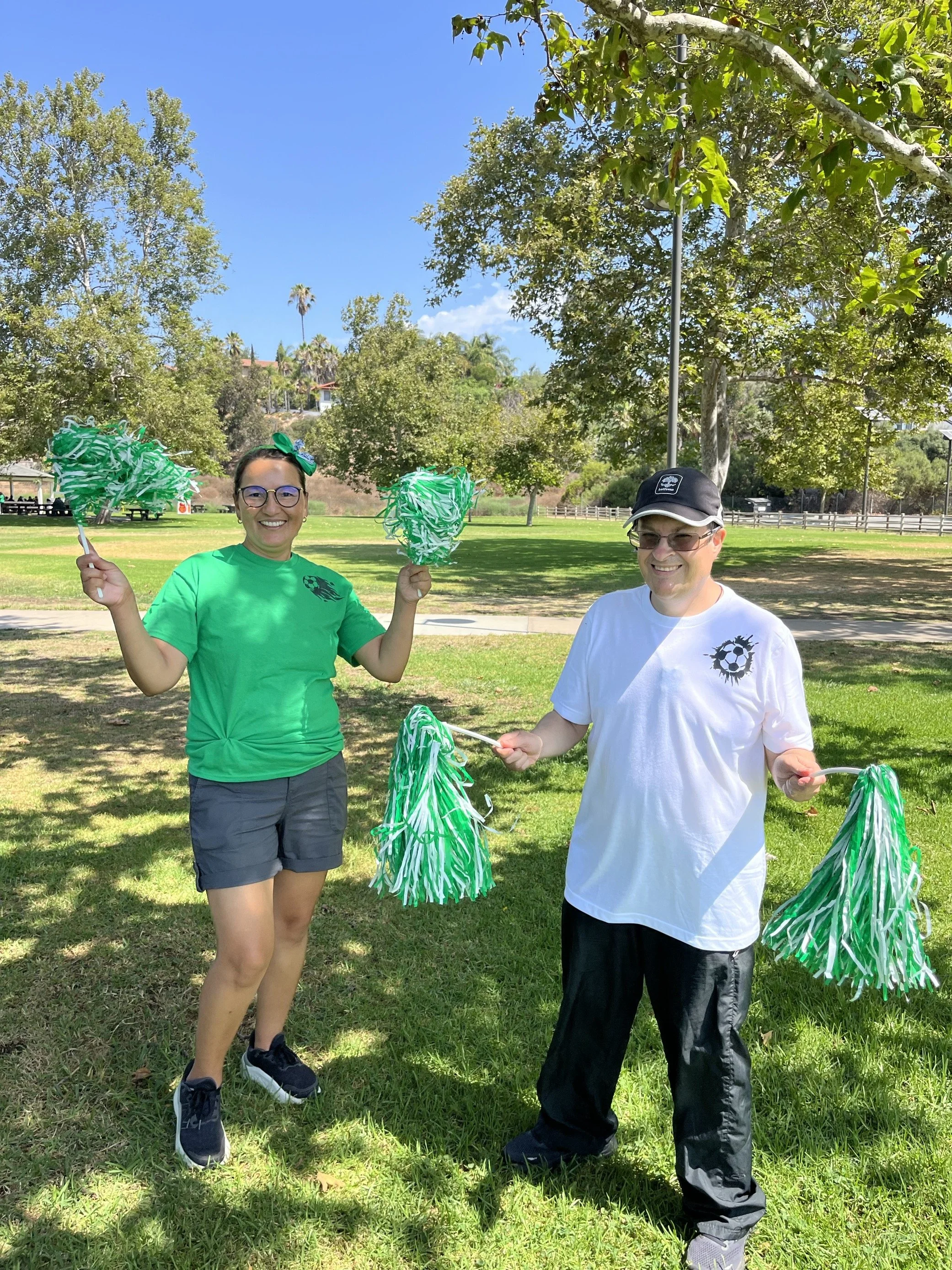 Two people standing outdoors on grass, smiling and holding green and white pom-poms, with trees and a park in the background on a sunny day.