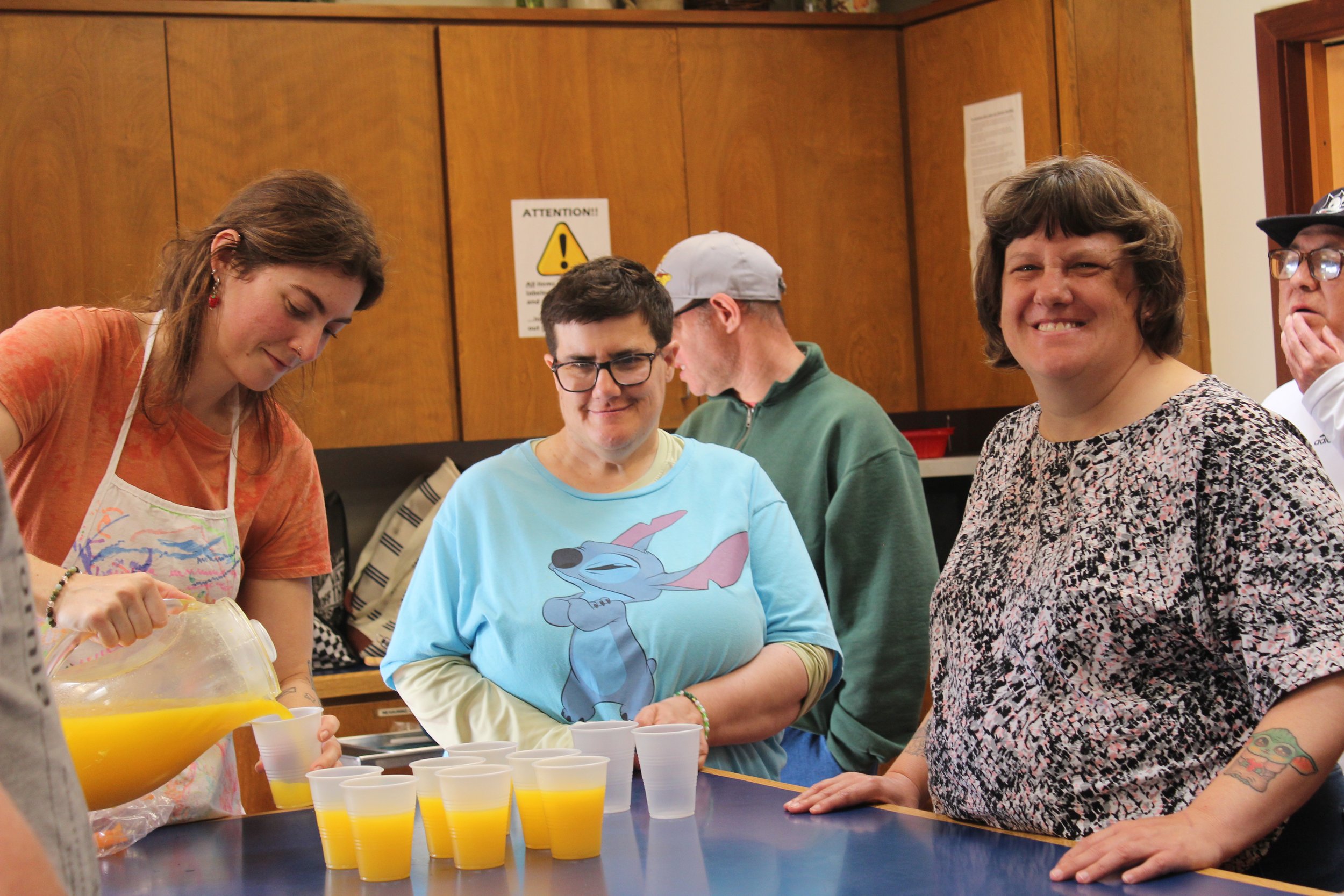 A group of clients standing around a table with cups of orange juice, some of them smiling and engaging in conversation in a room with wooden paneling.