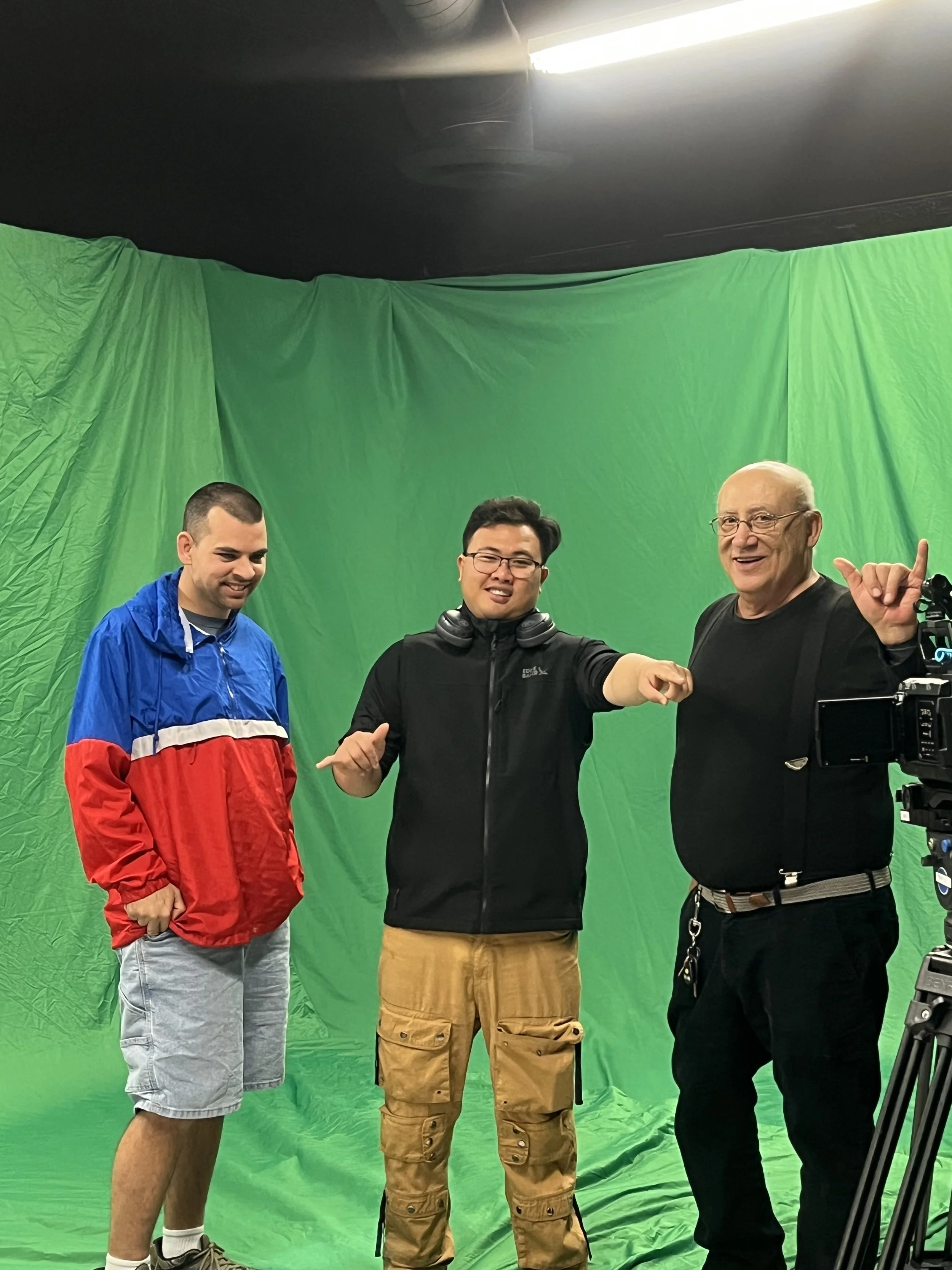 Three men standing in front of a green screen, smiling and posing for a photo. The man on the left is wearing a red, blue, and white jacket with gray shorts. The man in the middle is wearing a black jacket and khaki cargo pants, with headphones around his neck. The man on the right is wearing a black shirt and glasses, making a gesture with his right hand.