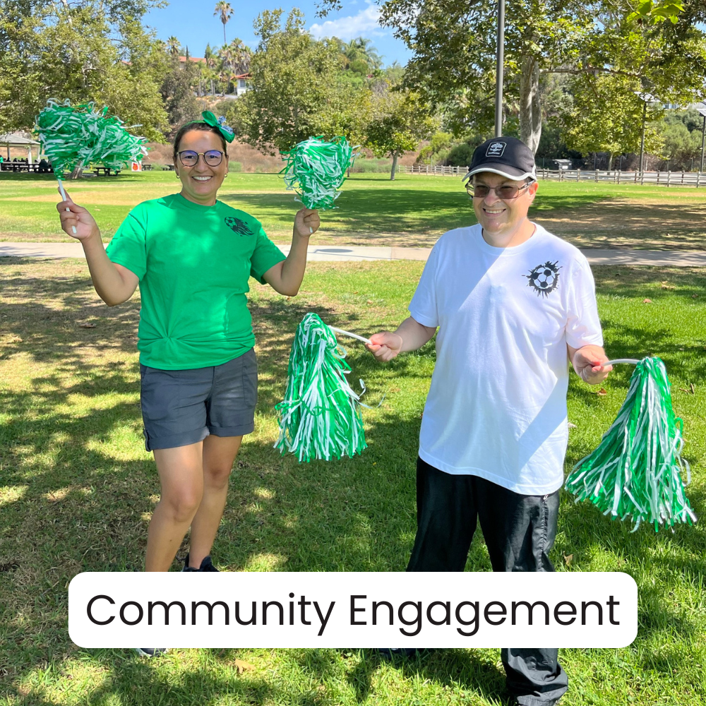 Two people smiling and holding green and white pom-poms, standing on grass in a park with trees and a fence in the background, participating in community engagement activity.
