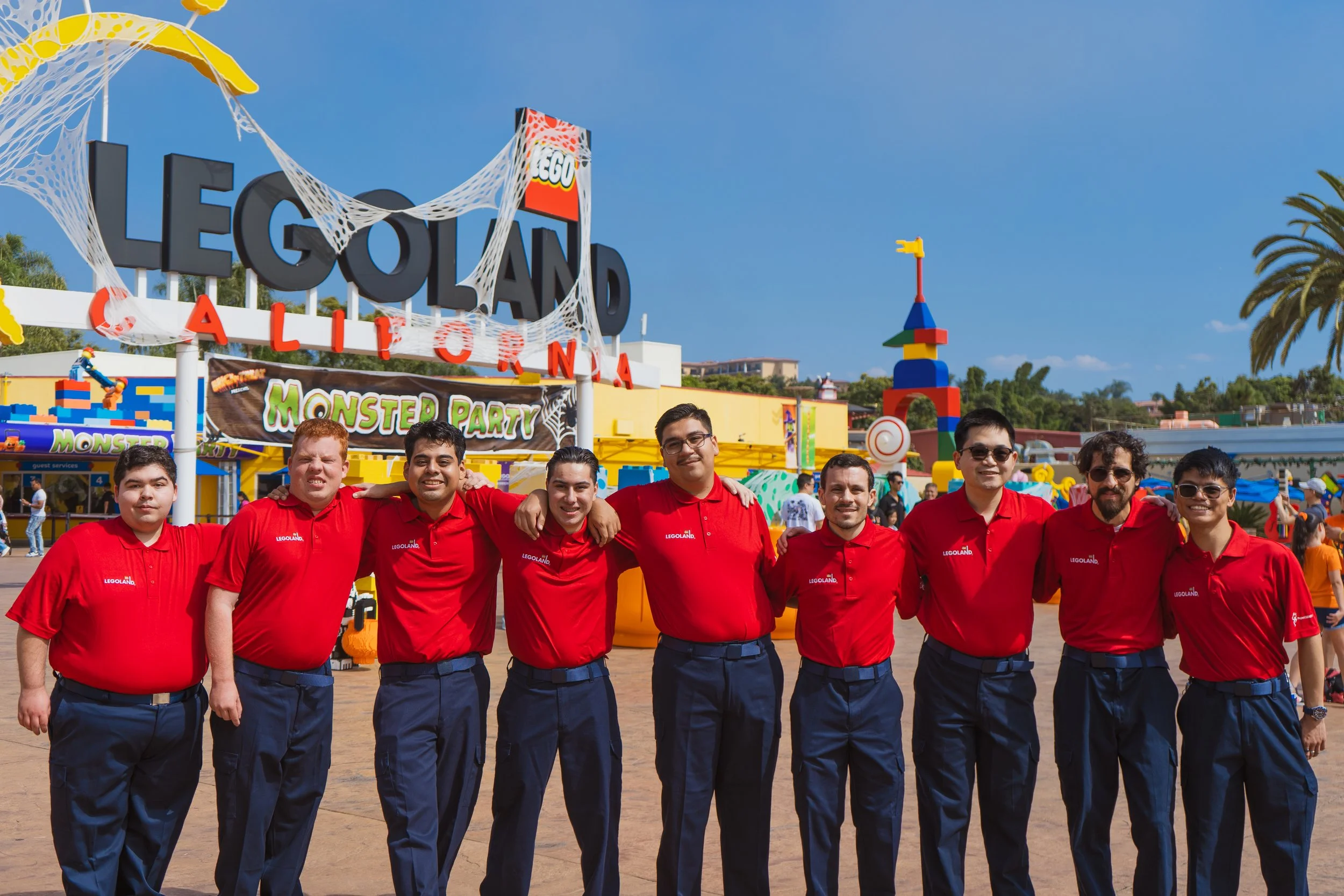 A group of seven employees in red shirts standing arm-in-arm in front of LEGOLAND California entrance with colorful LEGO structures and sign, under a clear blue sky.